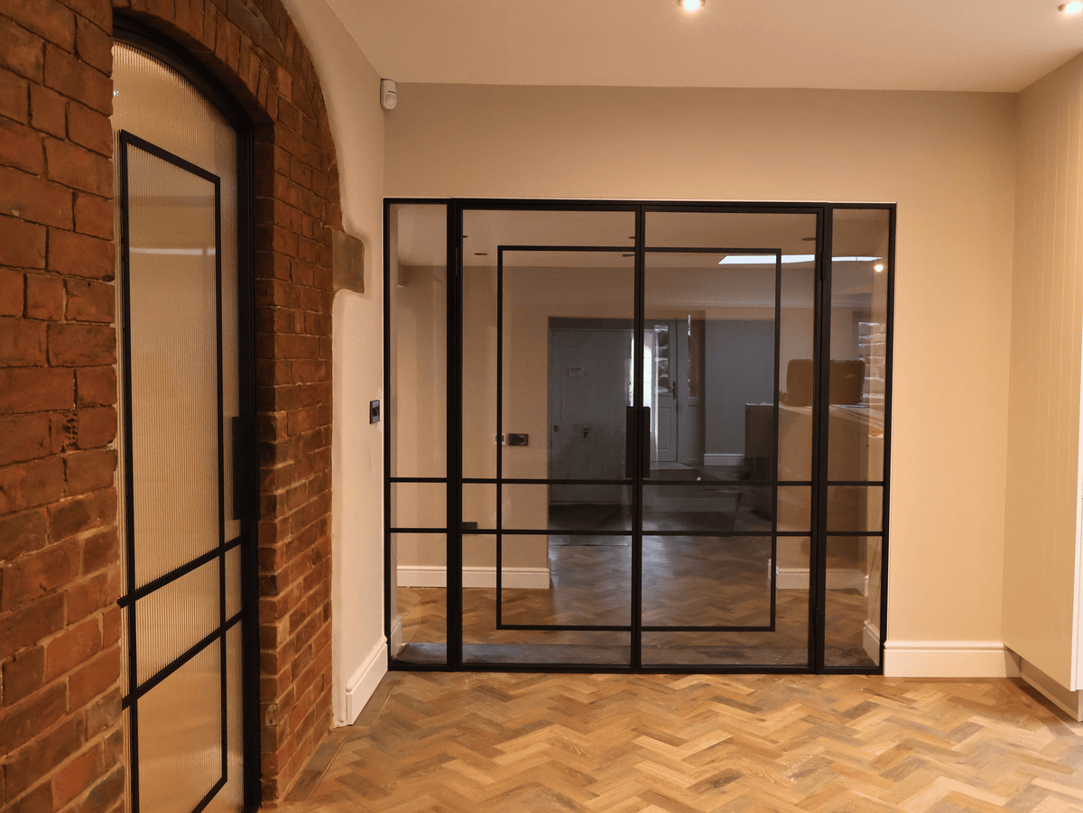 Modern glass partition and arched single door in a room with brick wall and wooden floor.