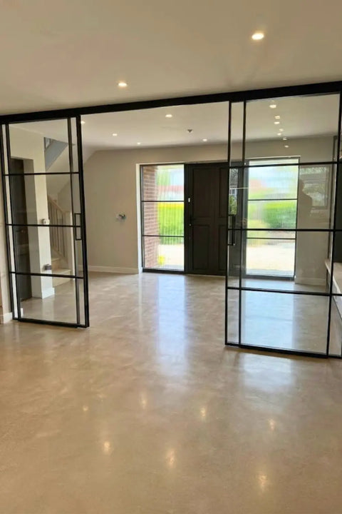 Modern interior space with polished light beige stone flooring, black-framed glass sliding doors, and large windows looking out onto a garden.