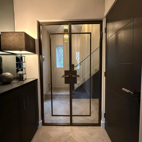 A modern double steel and glass door with a dark metal frame and handle, installed in an entryway next to a dark wood door.