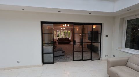 Half-open black steel concertina sliding door leading into a bright conservatory, featuring slimline steel frames and glazed panels allowing natural light through the space.