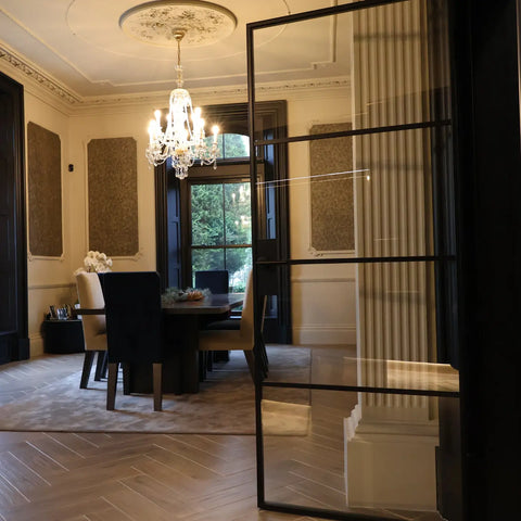 A modern black-framed glass door with a vertical slatted design, shown in a contemporary dining room setting with a chandelier, wooden floors, and a dining table.